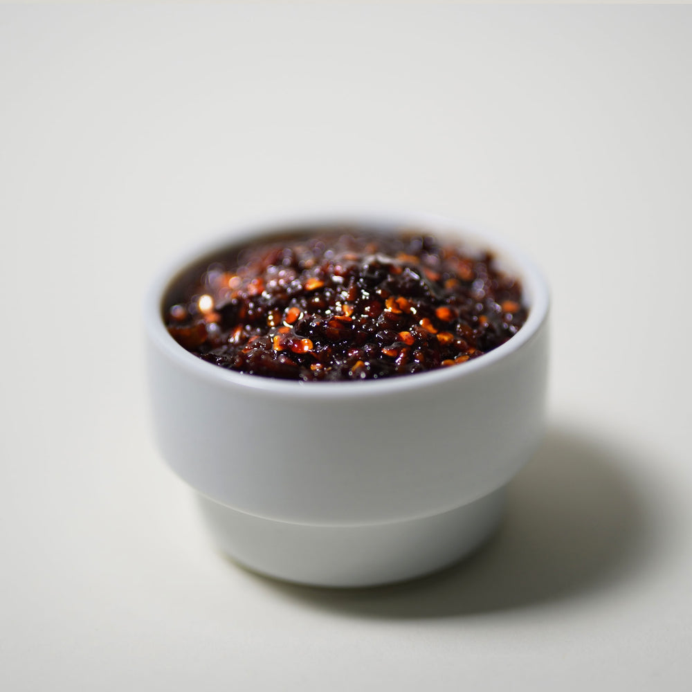 Close-up image of a white ceramic cup filled with vibrant red Thai chili paste, set against a neutral background to highlight the rich color and texture of the paste. The chili paste appears thick and glossy, with visible bits of chili and herbs, suggesting its freshness and homemade quality.